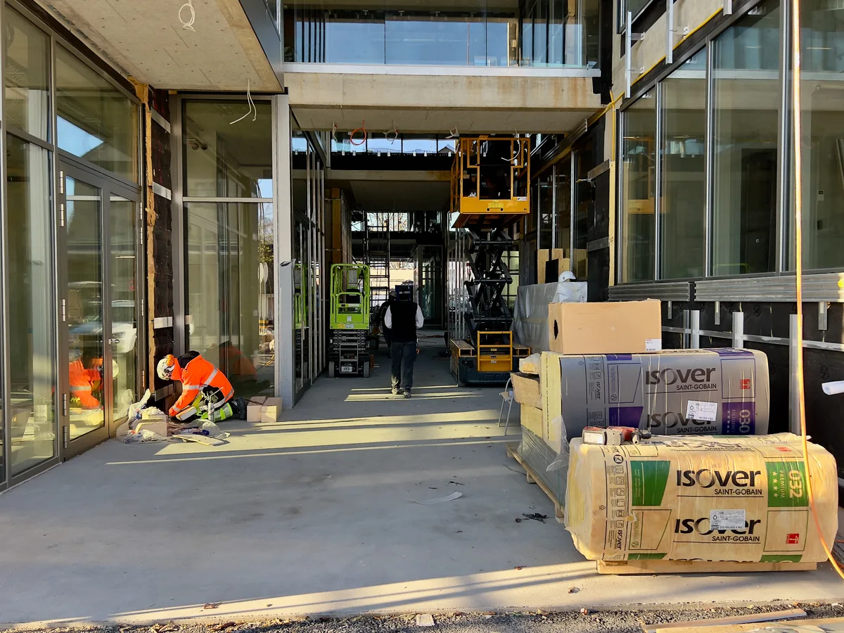 Im Bild ist ein Durchgang zu sehen, der links und rechts von den Fensterfronten des Gebäudes gesäumt wird. Links ist ein Handwerker damit beschäftigt Dämmmaterial zu montieren. Rechts steht noch ein Haufen Dämmmaterial bereit.